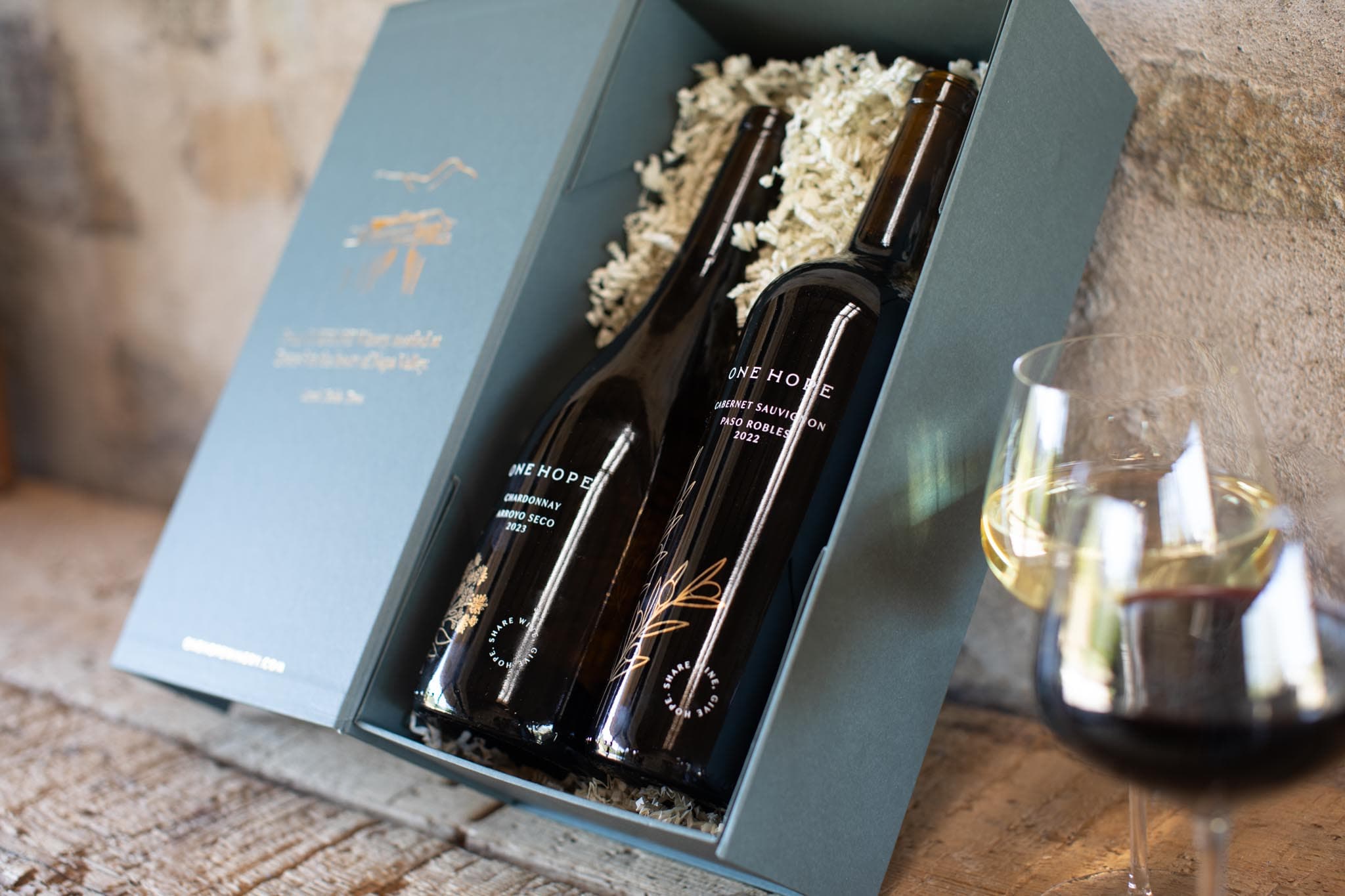 Two ONEHOPE wine bottles presented in an open green gift box beside wine glasses